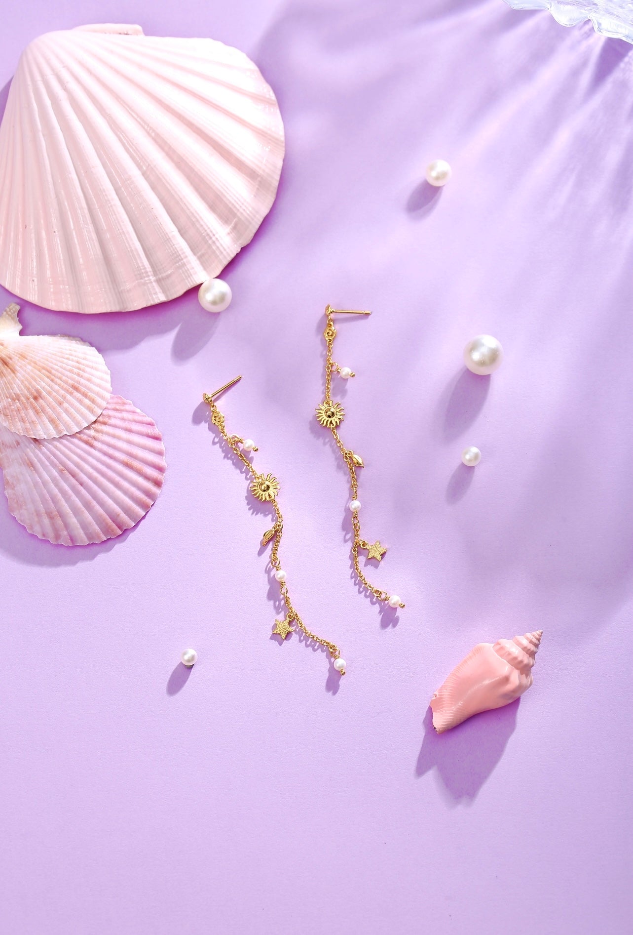 Pallantis Smykker Drømme Stjerne Øreringe i forgyldt sølv med sol og stjerne charms og ferskvandsperler er stylet på en pastel lilla overflade med lyserøde muslingeskaller og perler, hvilket skaber en elegant beachy vibe.