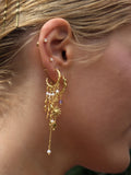 Nærbillede af en persons øre iført Pallantis Smykker Lilla Sol Øreringe, forgyldt sølv bøjler med dinglende charms og ferskvandsperler, sammen med mindre guld- og perleknopper. Blondt hår er delvist synligt.