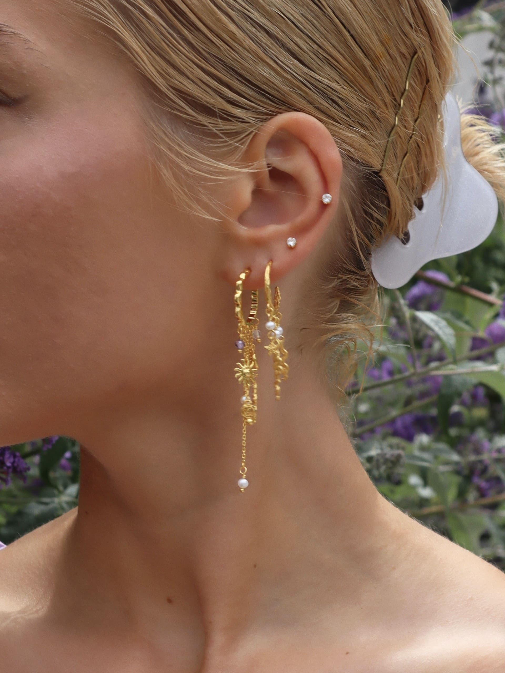 Et nærbillede af en kvindes øre viser to små ørestikker og elegante guldfarvede Lilla Sol-øreringe fra Pallantis Smykker. Hendes blonde hår er trukket tilbage med en hvid klemme, og bløde lilla blomster slører sig i baggrunden.