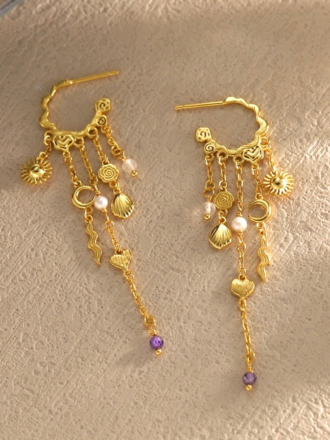 Pallantis Ocean Heart Øreringe er udsmykkede guldøreringe i forgyldt sølv med sole, måner, hjerter, muslingeskaller, ferskvandsperler, bølgede stænger og lilla perler hængende fra en dekorativ buet bøjle.