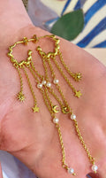 En hånd holder Pallantis Månestjerne Øreringe - guldøreringe med dinglende kæder, perler og charms formet som sole, stjerner og måner. Smukke himmelske smykker til enhver anledning.