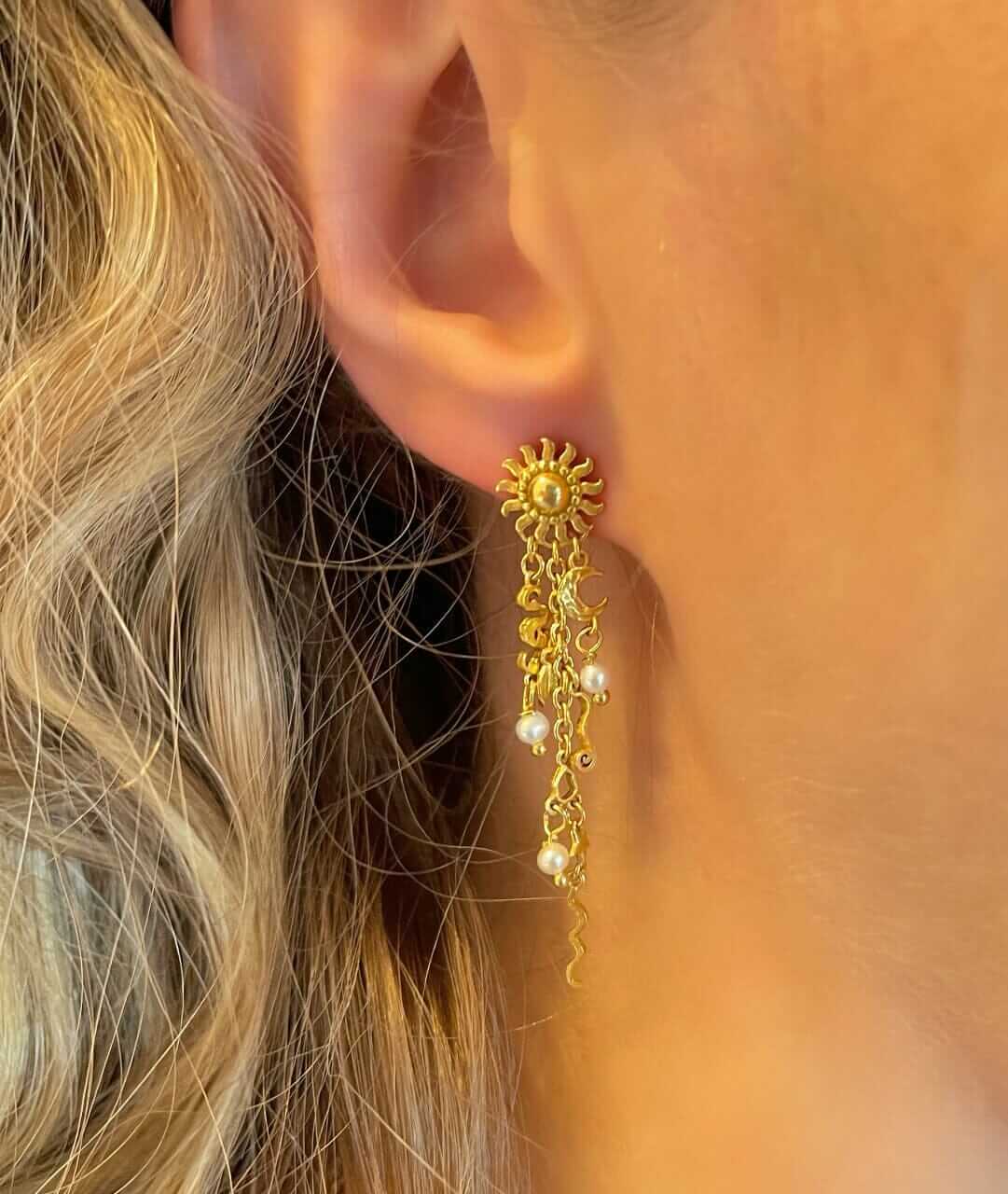 Nærbillede af Pallantis Sol Drømme Øreringe i forgyldt sølv, formet som en sol med dinglende kæder og små hvide ferskvandsperler, båret på et øre med delvist synligt bølget blond hår.