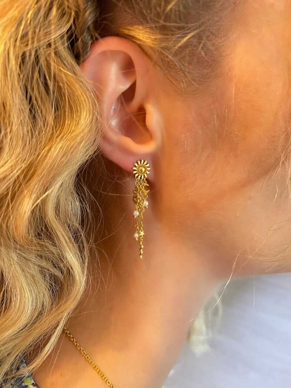Nærbillede af en persons øre med krøllet blond hår iført Pallantis Sol Drømme Øreringe – forgyldte øreringe med solmotiv og fine kæder med ferskvandsperler.