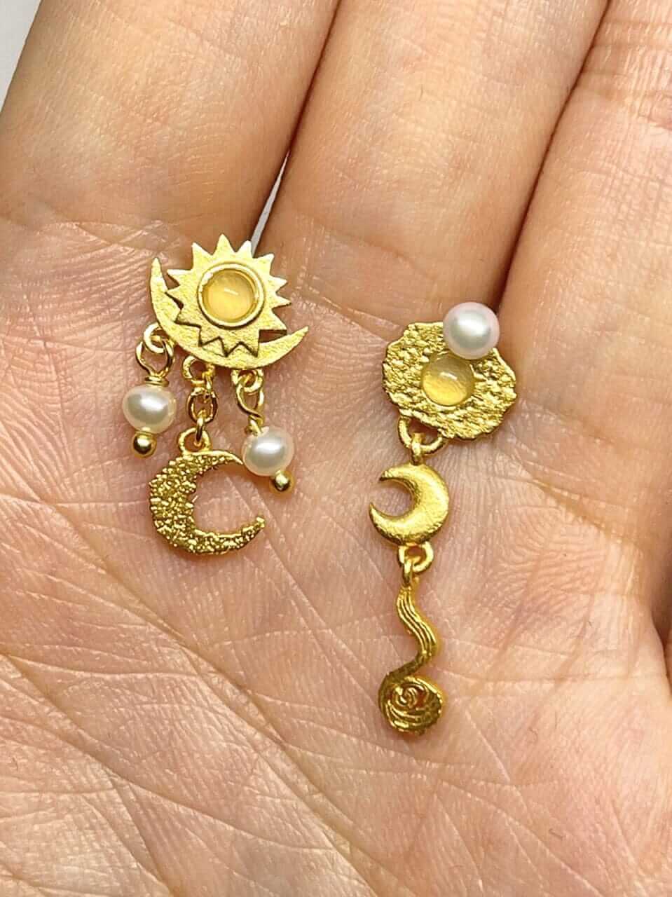 To Pallantis Sol Øreringe Med Månesten hviler på en håndflade. Den ene har sol, måne og perler; den anden har en tekstureret cirkel, halvmåne, månesten og bølget dråbe – begge inspireret af himmelske og naturlige motiver.